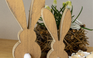 Zwei Osterhasen aus Holz stehen auf einem Tisch vor einem Frühlingsblüher im Korb.