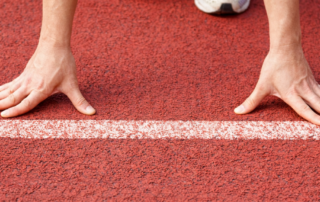 Zwei Hände an einer Startlinie einer Aschbahn.