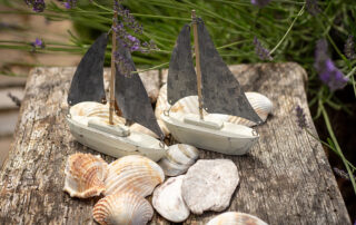 Zwei Miniatur-Segelbote aus Holz stehen zur Dekoration auf einem Tisch, Muscheln liegen daneben.