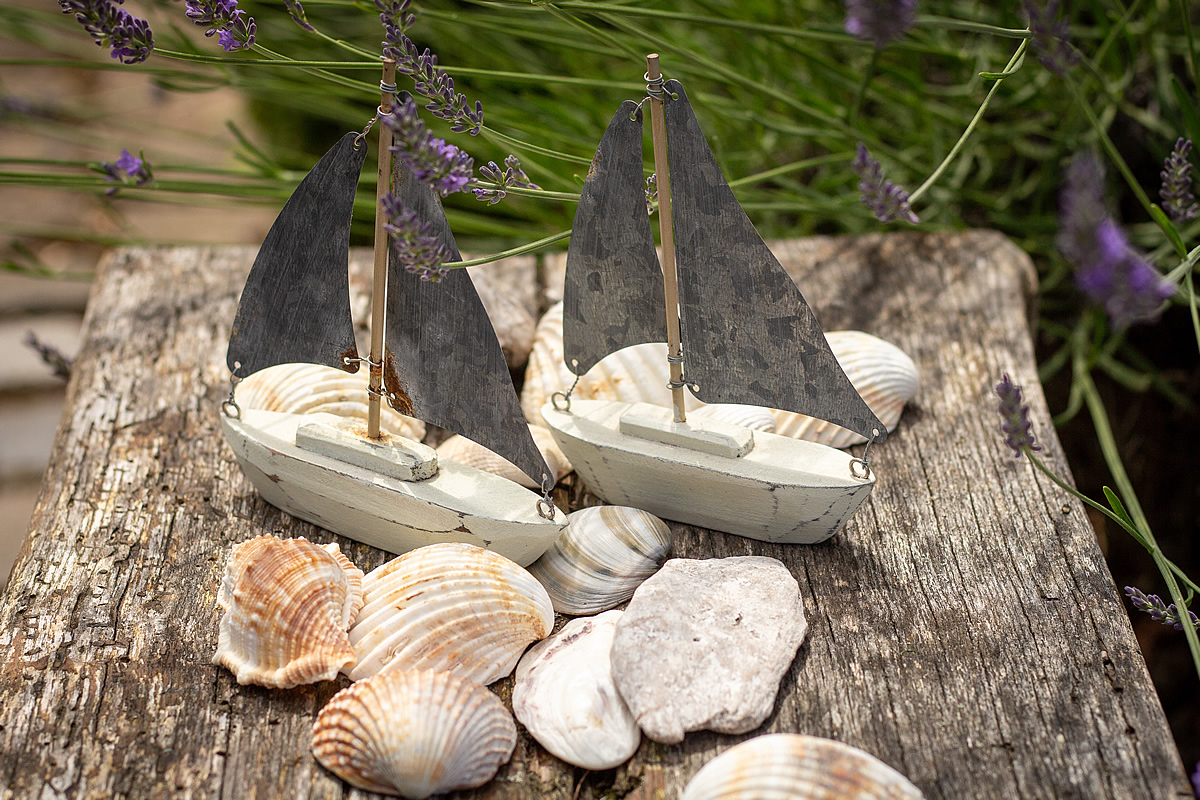Zwei Miniatur-Segelbote aus Holz stehen zur Dekoration auf einem Tisch, Muscheln liegen daneben.