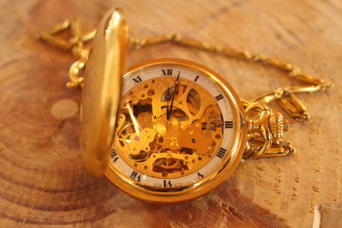 Eine goldene Taschenuhr liegt aufgeklappt auf einem Tisch und das Ziffernblatt sowie die dahinterliegende Mechanik sind zu sehen.
