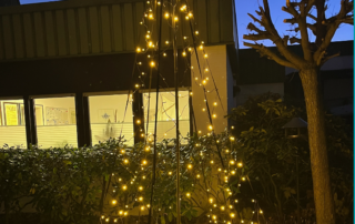 Ein Baum aus Lichtern steht in der Dunkelheit vor einem Haus.