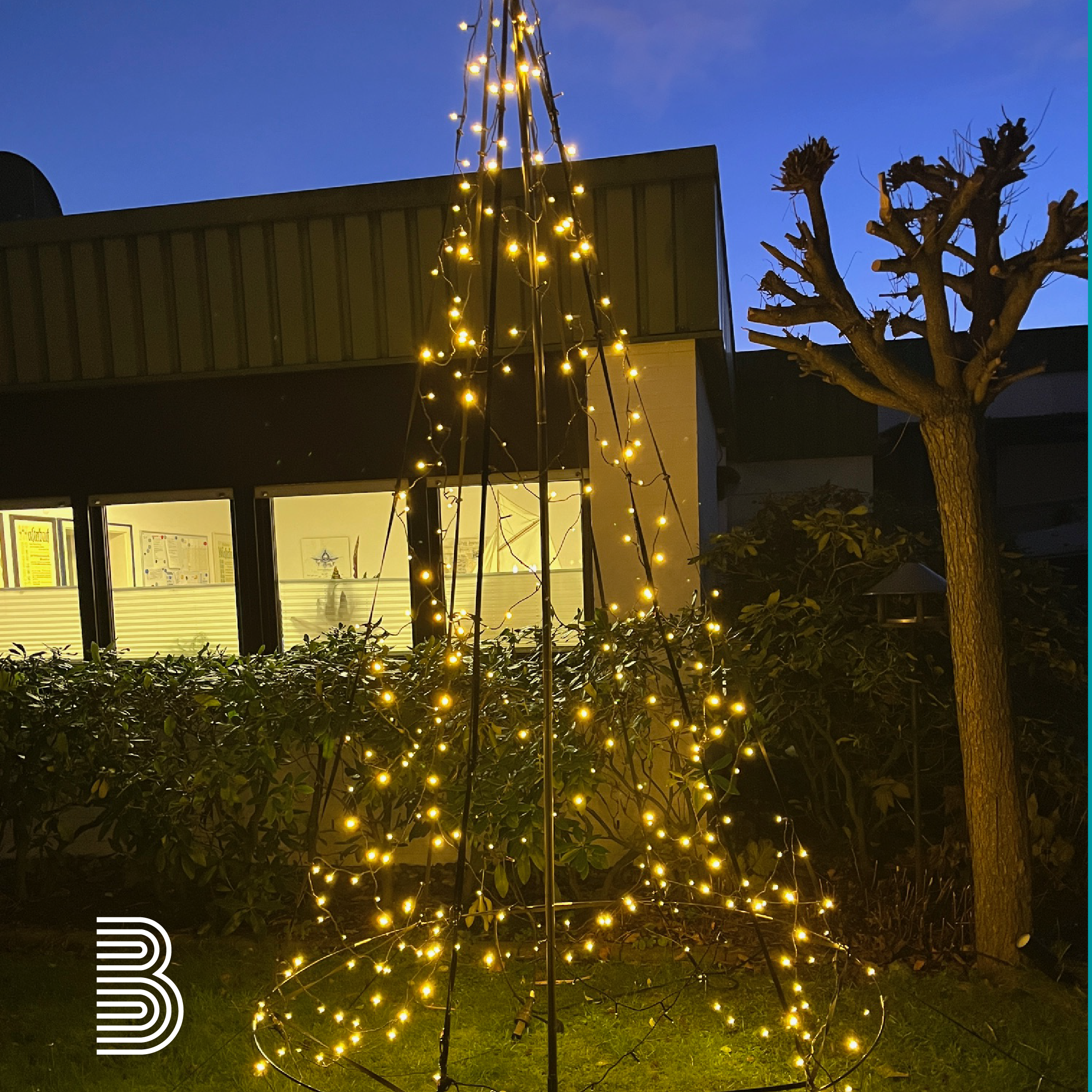 Ein Baum aus Lichtern steht in der Dunkelheit vor einem Haus.